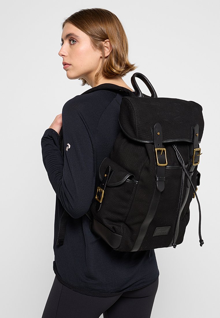 Schwarzer Canvas-Rucksack mit Lederakzenten, gerolltem oberen Teil, goldenen Beschlägen und verstellbaren Trägern, auf dem Rücken über einem dunklen Oberteil getragen.