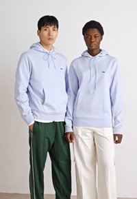 Hoodies lilás claros com bolsos canguru, combinados com calças listradas verdes e brancas à esquerda, e calças cargo brancas à direita.