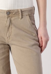 Beige bavlněné kalhoty mají vysoký pas, postranní kapsy a hladký povrch. Knoflíkové zapínání je kovové s kontrastním šitím.