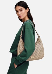 Borsa trapuntata beige con dettaglio logo, dotata di una singola tracolla. Indossata con una felpa verde e pantaloni della tuta abbinati.