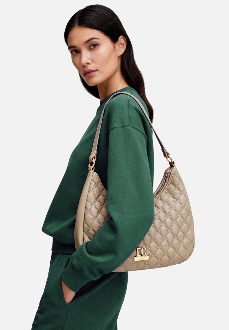 Borsa trapuntata beige con dettaglio logo, dotata di una singola tracolla. Indossata con una felpa verde e pantaloni della tuta abbinati.
