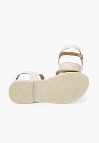 White sandals with a brown accent strap and a textured beige sole featuring a star pattern. Adjustable buckle closure on the back.