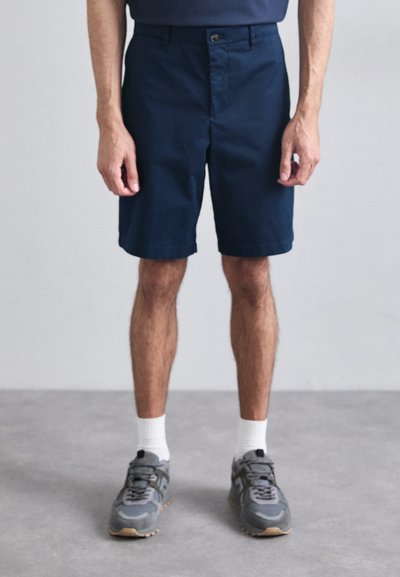 Marineblå shorts lavet af bomuldsblanding med flad front, bæltestropper og enkelt knaplukning. Båret med grå sneakers og hvide ankelstrømper.