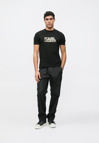 Čierne bavlnené tričko so zlatým logom "KARL LAGERFELD", doplnené o čierne nohavice a čierne tenisky, na modeli na jednoliatom pozadí.