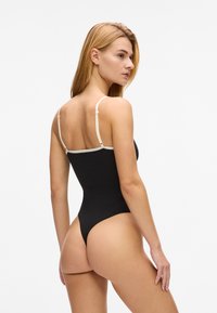 Crni kupaći kostim s tankim bež naramenicama, visokog izreza i minimalnim pokrivanjem na leđima. Glatka tekstura, prilagođen kroj.