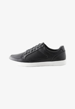 Zapatilla deportiva negra de corte bajo con suela blanca, cordones en el frontal y panel lateral de malla, mostrada desde el lado exterior sobre un fondo blanco.