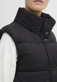 Schwarze gepolsterte Weste mit hohem Kragen, versehen mit Druckknopfverschlüssen und seitlichen Schlitzen. Der Stoff wirkt glatt mit einer glänzenden Oberfläche.