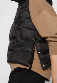 Gilet imbottito nero con finitura testurizzata, dotato di colletto alto, bottoni laterali e design stratificato sopra un sudario marrone.