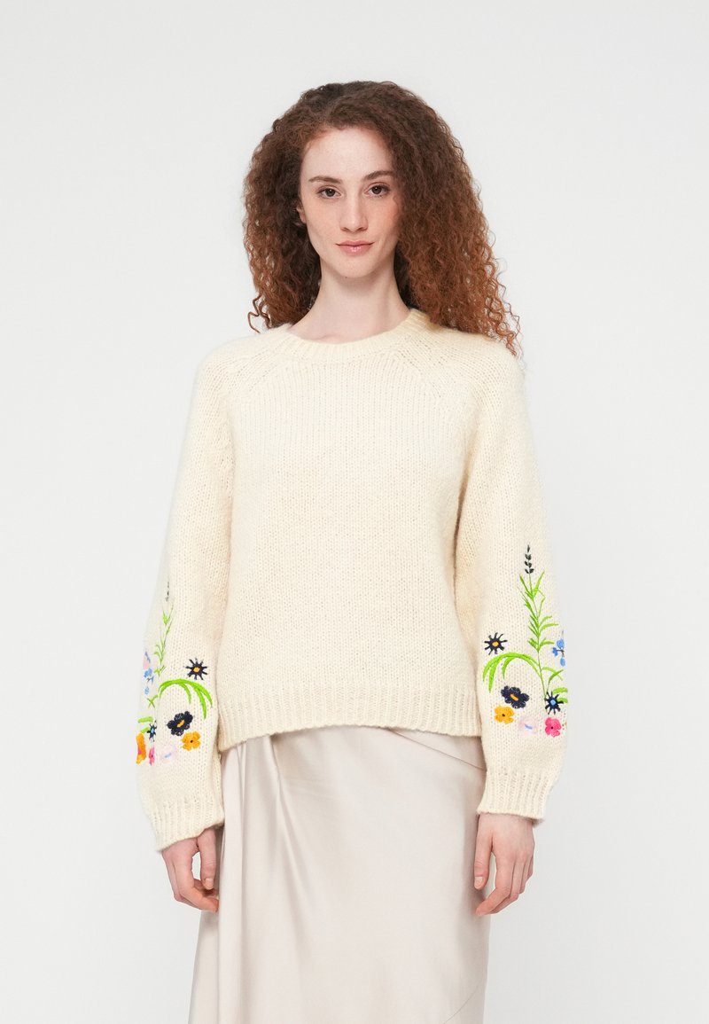 Femme aux cheveux bouclés portant un pull crème avec une broderie florale colorée sur les manches et une jupe beige, debout devant un fond blanc.