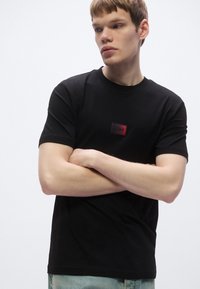 Μαύρο βαμβακερό t-shirt με στρογγυλή λαιμόκοψη, κοντά μανίκια, με μικρό ορθογώνιο λογότυπο σε κόκκινο και μαύρο στο στήθος.