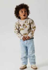 Beige sweatshirt met cartoon hondenprint, geribde boorden, gecombineerd met lichtblauwe baggy jeans en tan sneakers. Kind staat met handen op de heupen.