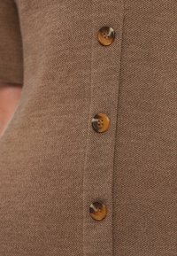 Καφέ πλεκτό τοπ με κοντά μανίκια και σειρά από στρογγυλά κουμπιά με τελείωμα σε σχέδιο χελώνας κατά μήκος της εμπρόσθιας πατιλέτας. Μαλακή υφή.