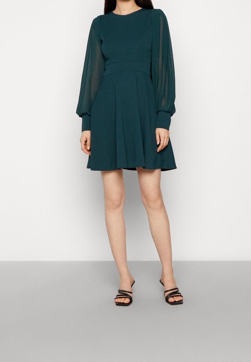 Femme portant une robe longue à manches longues vert foncé avec des manches transparentes et des sandales à talons noires à lanières, debout sur un sol gris.
