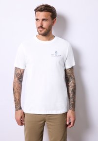 Bílý bavlněný tričko s malou grafikou v modré barvě na levé části hrudi, spárované s béžovými kalhotami. Texturované manžety a hladký lem.