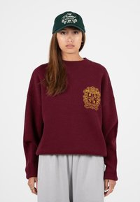 Burgundowy, oversize sweter z teksturowanym, złotym herbem. Szare, luźne spodnie i zielona czapka baseballowa z białym napisem.
