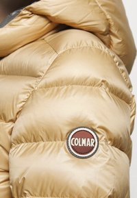Gouden pufferjacksectie met glanzende, doorgestikte textuur; heeft een ronde "COLMAR" logo patch in bruin en wit aan de voorkant.