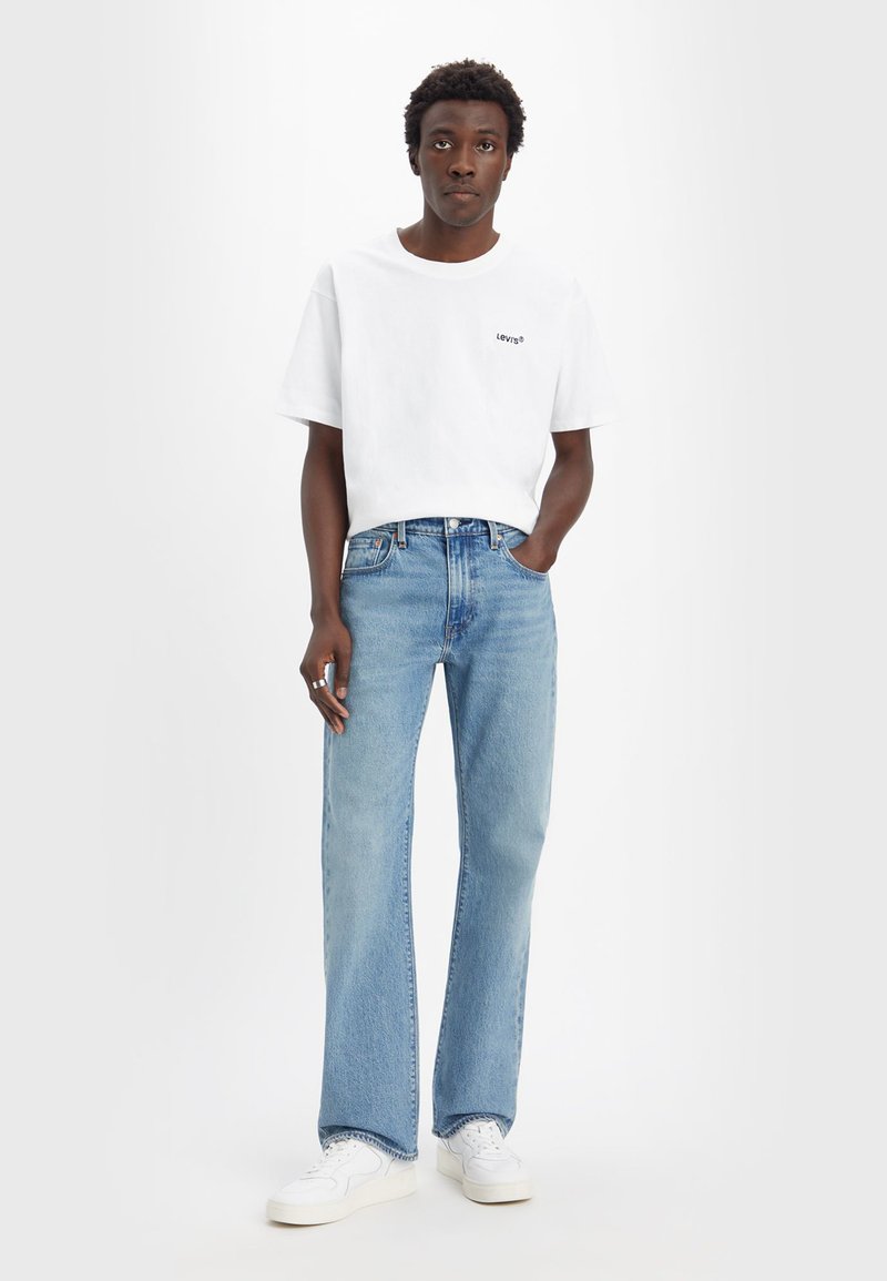 Weißes Baumwoll-T-Shirt mit kleinem schwarzem Logo, kombiniert mit hellblauen Denim-Jeans, im geraden Schnitt, und weißen Sneakers.