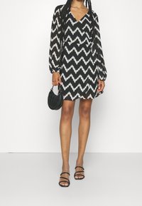 Svart och vit klänning med zigzag-mönster, långa ärmar, v-ringning och utsvängd nederdel, i kombination med en liten svart handväska och sandaler med remmar.