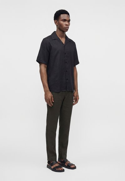 Homme debout portant une chemise noire à manches courtes boutonnée, un pantalon olive foncé et des sandales noires ouvertes aux orteils sur un fond clair uni.