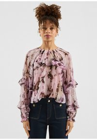 Transparante blouse met gerimpelde halslijn en mouwen, met een abstract patroon in roze en bruin. Gecombineerd met donkere denimbroek.