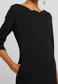 Černé šaty s vroubkovaným výstřihem, přiléhavým střihem, tříčtvrtečními rukávy a bočními kapsami. Vyrobeno z strukturované tkaniny.