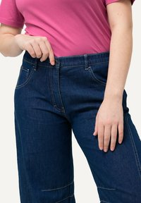 Dunkelblaue Denim-Hose mit breitem Bund, frontalen Taschen und sichtbaren Nähten. Das Modell trägt ein pinkes T-Shirt und passt den Bund an.