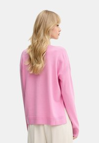 Donna con lunghi capelli biondi che indossa un maglione rosa largo e pantaloni beige chiari, rivolta di lato contro uno sfondo chiaro e semplice.
