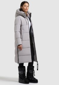 Lång ljusgrå pufferjacka med huva och horisontella sömmar, kombinerad med svarta snöboots med snörning och logotyp.