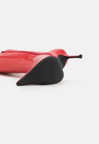 Red leather high heel shoe with a pointed toe and a sleek silhouette. Features a zip closure and a black rubber sole. Smooth texture.