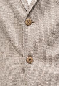 Μπεζ υφασμάτινο σακάκι με δύο καφέ κουμπιά, το ένα κουμπωμένο στο στήθος και το άλλο στη μέση, με λεπτομέρεια του υφάσματος σε κοντινό πλάνο.