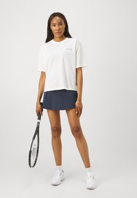 Top alb, din plasă, cu mânecă scurtă și guler rotund. Asortat cu o fustă albastru închis și ținând o rachetă de tenis neagră. Adidașii albi completează ținuta.