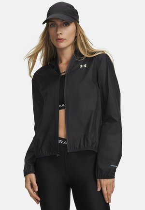 Frau mit schwarzer Mütze, schwarzer Zip-Jacke mit Logo und schwarzen Sportleggings vor einem einfarbigen hellen Hintergrund.