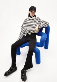 Maglione a righe bianche e nere, pantaloni neri a vita alta, sandali neri e un cappellino nero, seduto su una sedia scultorea blu.