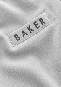 Biela tkanina s textúrovaným povrchom, na ktorom je našitý obdĺžnikový štítok s nápisom "BAKER" tmavošedými písmenami.