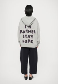 Grijze hoodie met grote donkere paarse tekst op de achterkant die "IK BLIJF LIEVER THUIS" zegt. Voorzien van een capuchon en een relaxte pasvorm, gecombineerd met donkere broeken.
