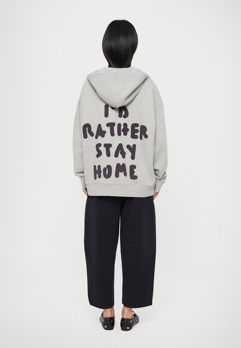Grijze hoodie met grote donkere paarse tekst op de achterkant die "IK BLIJF LIEVER THUIS" zegt. Voorzien van een capuchon en een relaxte pasvorm, gecombineerd met donkere broeken.
