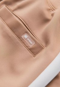 Beige trainingsbroeken hebben een soepele stof, een patchzak met een duidelijk label en contrasterende witte accenten langs de zijkant.