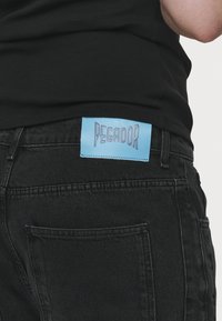 Personne portant un jean noir avec une étiquette de marque Pegador bleue cousue à la ceinture arrière, associée à un haut noir.