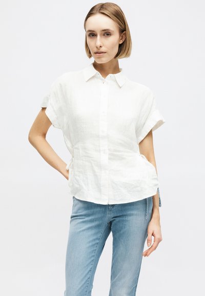 Femme aux cheveux blonds coupés au carré portant une chemise blanche à manches courtes et un jean bleu clair, debout devant un fond blanc uni.