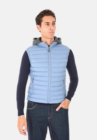 Uomo che indossa un gilet smanicato trapuntato azzurro chiaro sopra una maglietta a maniche lunghe scura e jeans scuri, in piedi con una mano in tasca.