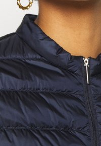 Close-up of person wearing a navy blue quilted jacket with a silver zipper and a gold hoop earring with round details.