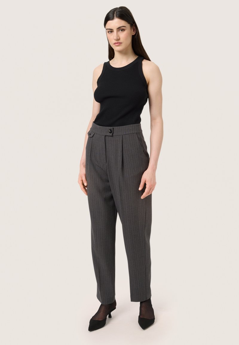 Un débardeur noir à côtes associé à un pantalon taille haute gris à rayures avec des plis avant et des jambes fuselées. Des escarpins noirs à talons pointus complètent le look.