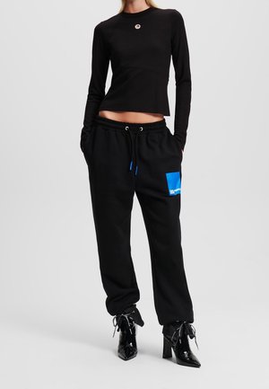 Vrouw draagt een zwart lang mouwcroptop, zwarte joggerbroek met een blauw patch en zwarte enkellaarsjes met hoge hakken, handen in de zakken.