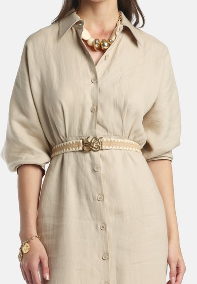 Kvinde iført beige knapklædt kjole med pufærmer, mønstret bælte med guldbeslag, chunky halskæde i guld og creme samt guldfarvet charme-armbånd.