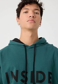 Sudadera color teal con cordones negros y la palabra "INSIDE" impresa en letras negras en negrita. Cuenta con una capucha negra de doble capa. Material de mezcla de algodón.