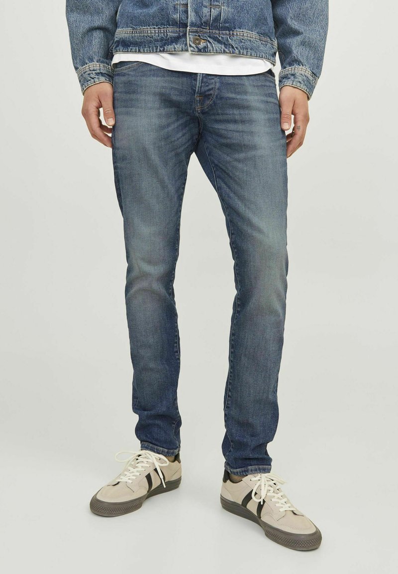Vaqueros de denim azul ajustados con un ligero desgastado, costuras cosidas y un sutil desvanecimiento. Combinados con zapatillas retro de color claro.