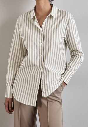 Persoon die een beige en wit verticaal gestreept overhemd met knopen draagt en beige broek, staand tegen een effen lichtgrijze muur.
