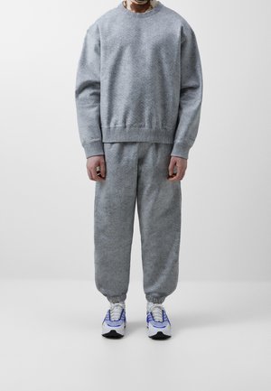 Mann trägt lässiges graues Sweatshirt mit Struktur und passende Jogginghose, dazu weiße und blaue Sneaker, steht vor einfachem weißen Hintergrund.