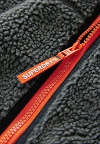 Giacca in pile grigia con una cerniera arancione brillante e un tirante, con un'etichetta tessuta che riporta "SUPERDRY®" vicino all'area della cerniera.