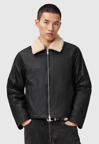 Chaqueta de cuero negra con un collar de borrego beige, cierre de cremallera, bolsillos laterales y un acabado texturizado. El diseño es contemporáneo y ajustado.